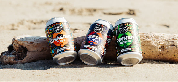 Three cans Sprig + Fern Brewing Co. 330ml cans of Chasing Hazy, Best Bitter and Pinner Pilsner on a beach with driftwood.
