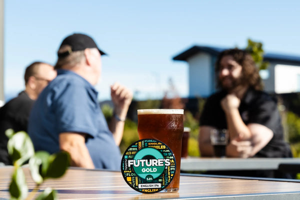 Glass of beer with 'Future's Gold' label on a table outdoors with people in the background at Sprig + Fern The Meadows
