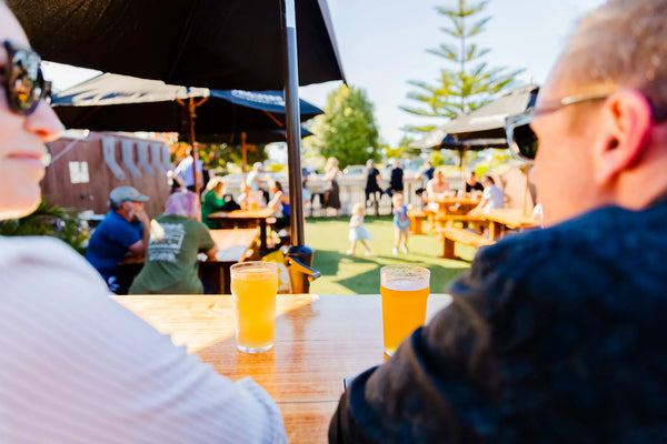 Two people sitting at an outdoor table with drinks, surrounded by a casual outdoor setting at Sprig + Fern Tahuna.
