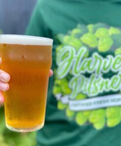 A pint of Sprig and Fern's fresh hop Harvest Pilsner being held by someone in a Harvest Pilsner tee shirt
