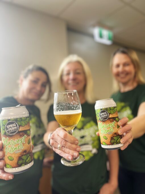 Three members of the Sprig and Fern team wearing Harvest Pilsner tee shirts and holding out cans and a glass of fresh hop Harvest Pilsner