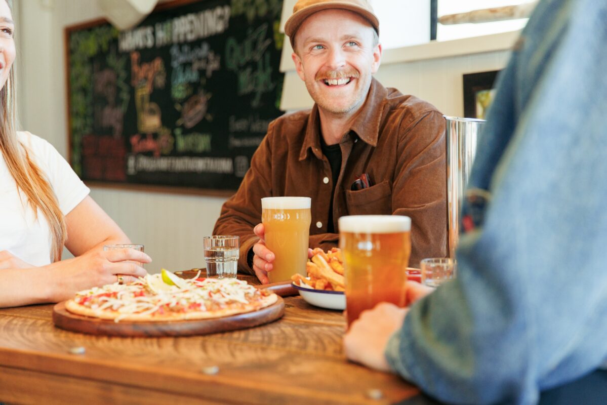 Three people sat around a table drinking beer and eating pizza.
